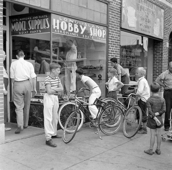 Group of youths on bicycles looking into the window of a Hobby Shop selling airplanes and train models. 